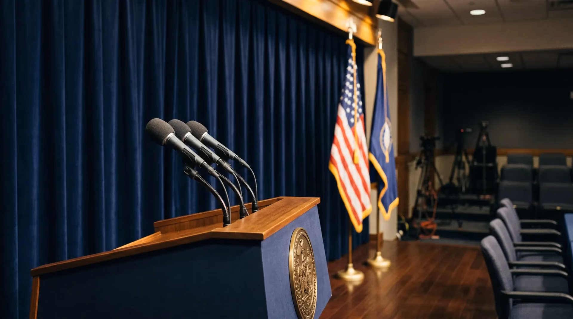 An empty press-conference podium with microphones in front of a navy curtain and flags